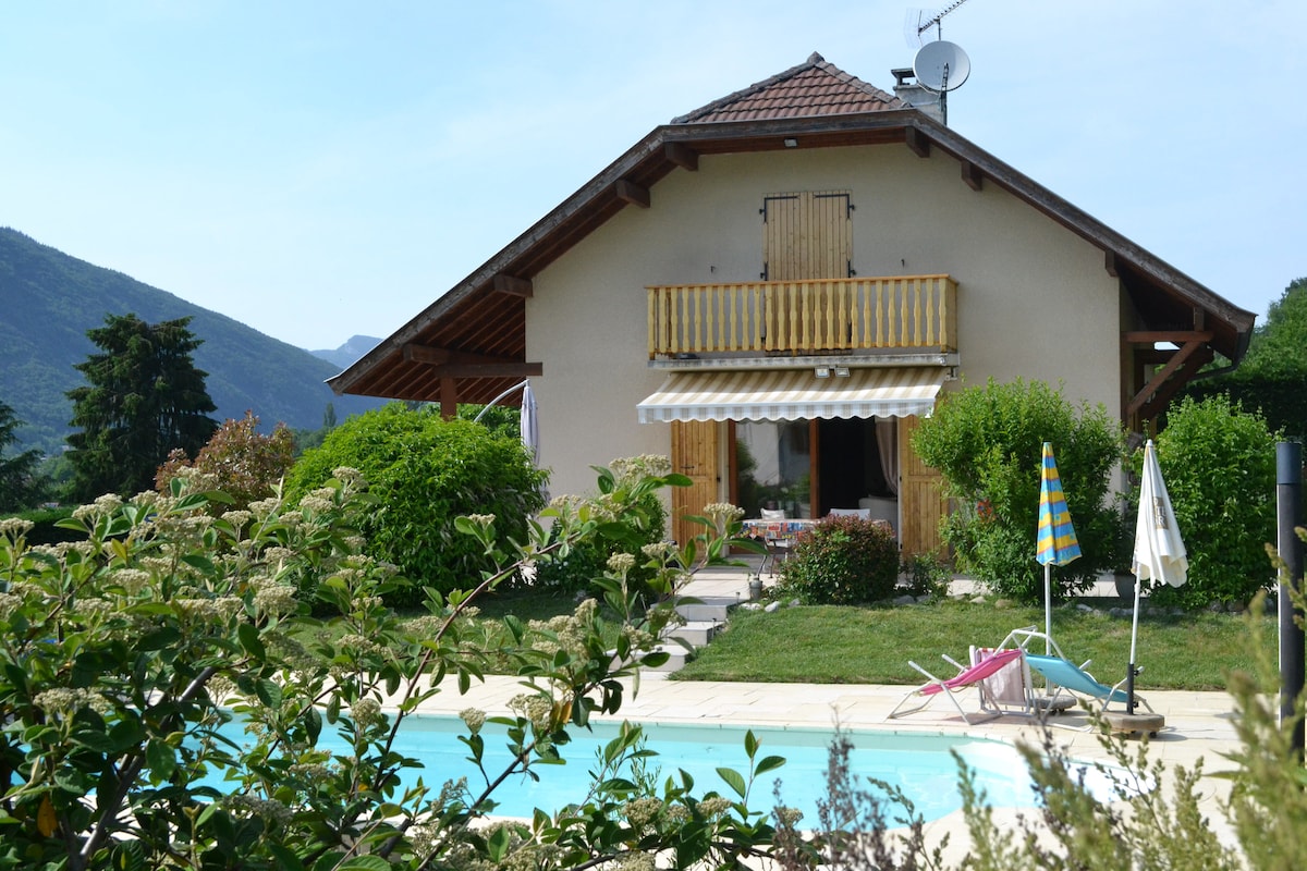 The house features a balcony with wooden railing, shaded by an awning. Lush greenery surrounds the structure, and a poolside area is visible, complete with lounge chairs and umbrellas. The backdrop includes gently sloping hills under a clear sky.