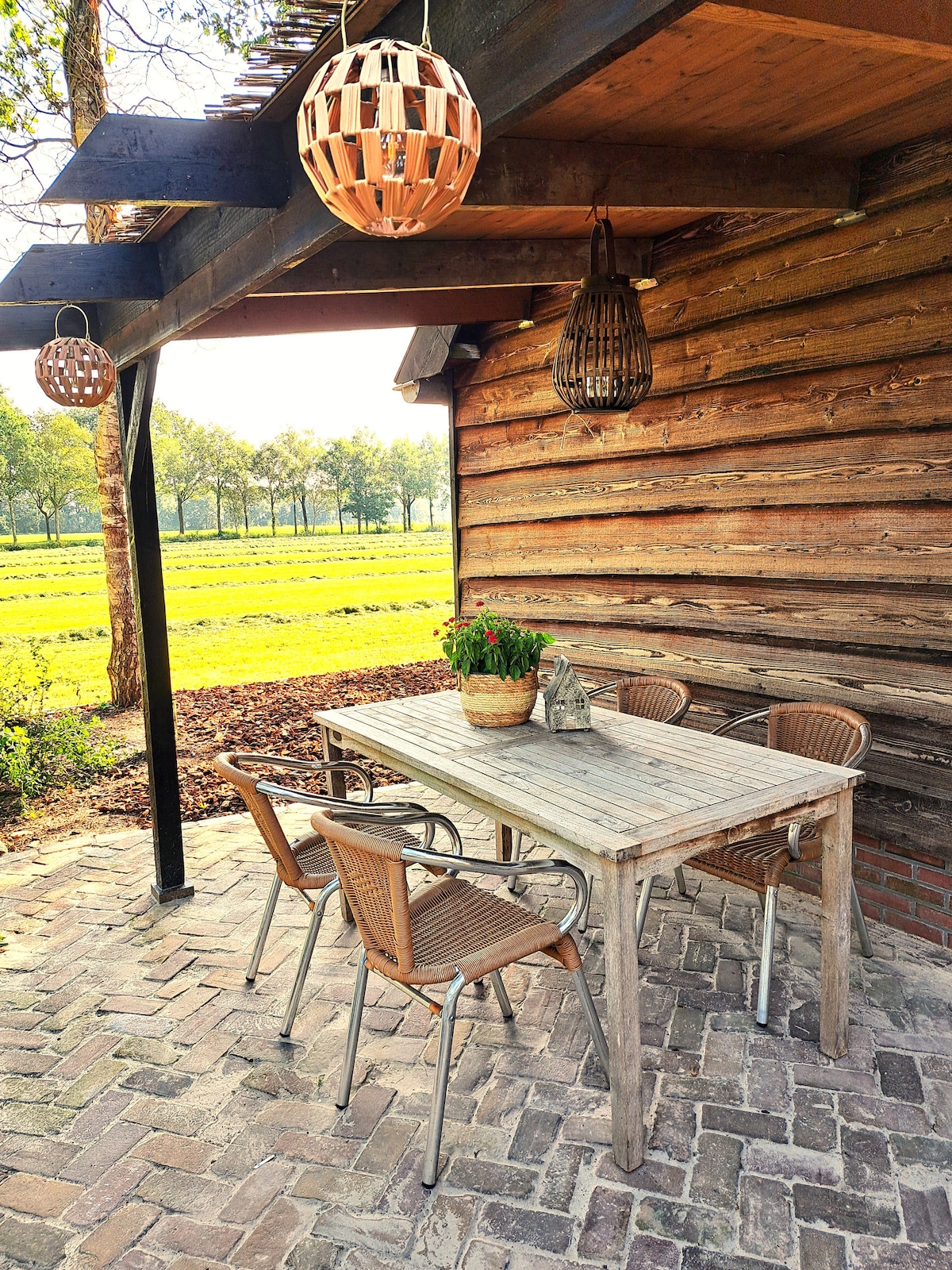 An outdoor dining area features a wooden table surrounded by four woven chairs. Hanging light fixtures add character above. A potted plant rests on the table, while a backdrop of lush green fields and trees is visible, creating a serene setting.