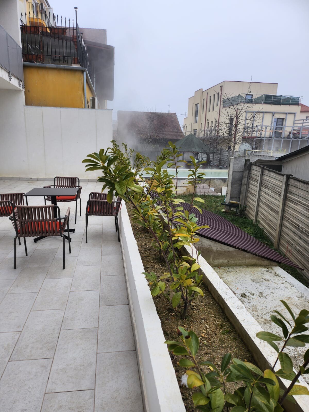 An outdoor terrace is presented, featuring several black chairs with red cushions around a small table. Alongside, neatly planted greenery adds an inviting touch. A view of an enclosed pool area is visible in the background, surrounded by various building structures.