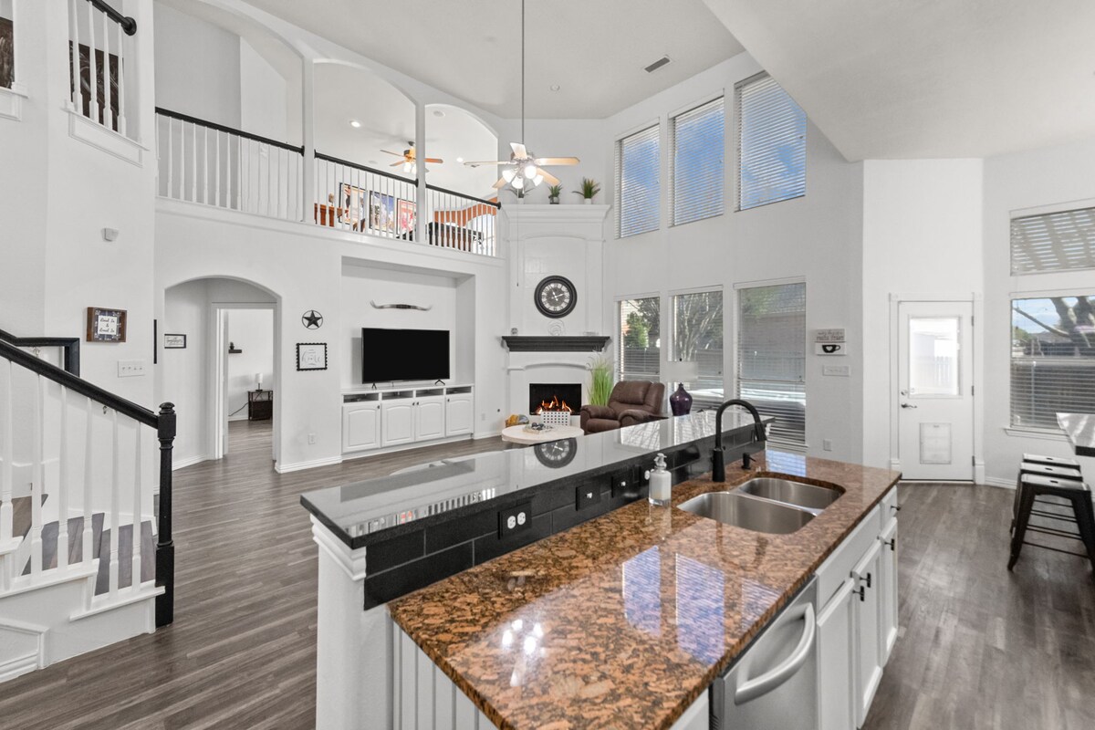 An open-concept kitchen features elegant granite countertops and a spacious island with a sink. The living area, visible in the background, is highlighted by large windows that allow natural light. White cabinetry complements the modern decor, and a comfortable seating area is seen beyond.