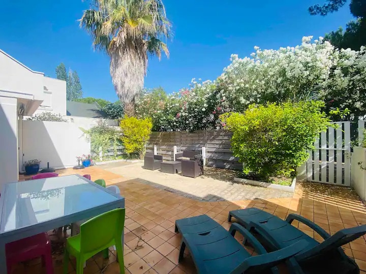 A well-maintained outdoor courtyard features sun loungers arranged on tiled flooring. Colorful chairs surround a dining table, while lush greenery and flowering plants create a serene environment. A wooden fence encloses the space, providing privacy and a tranquil atmosphere.