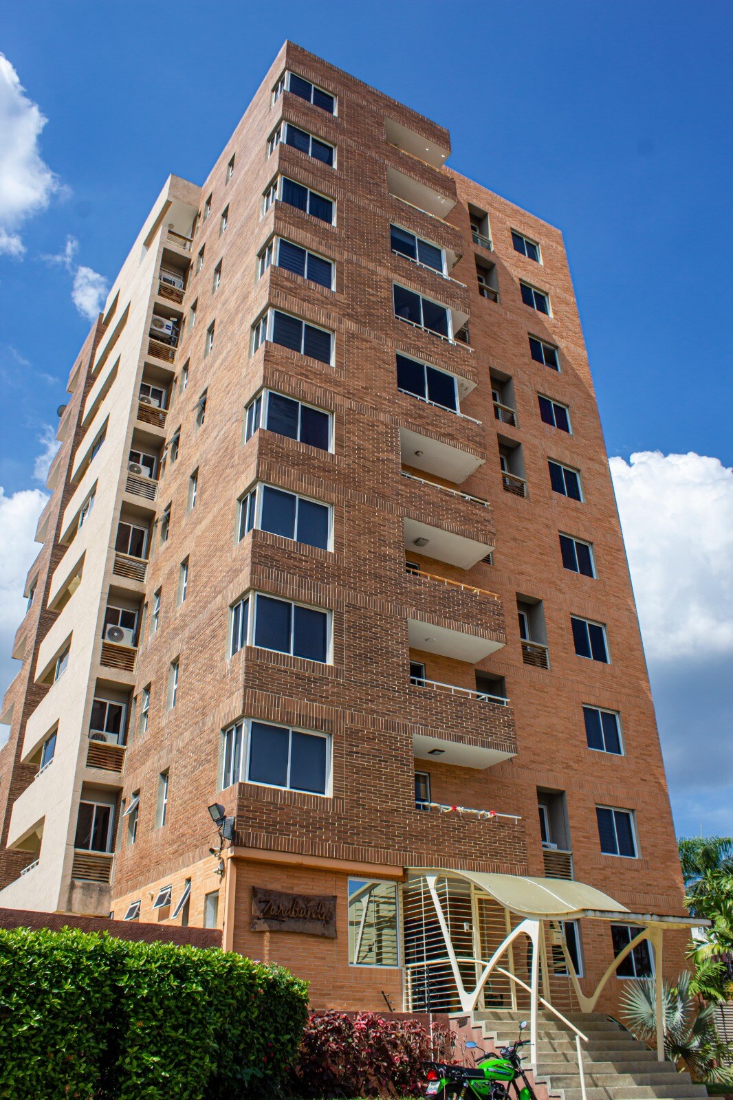 A modern multi-story building with a brick facade features numerous large windows, allowing ample natural light. A covered entrance leads to the lobby area. Balconies are visible on several floors, creating an inviting urban atmosphere.