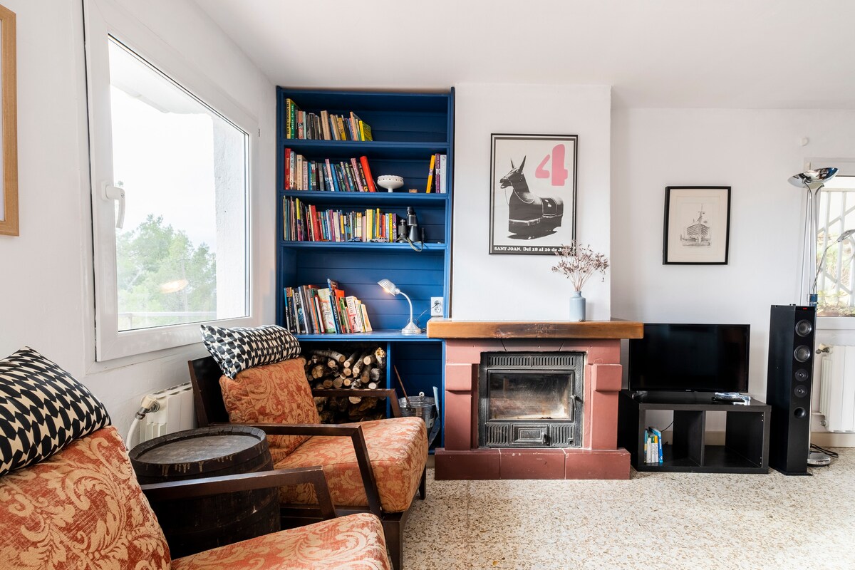 A cozy living area features two patterned armchairs positioned near a rustic fireplace. A built-in bookshelf filled with colorful books is complemented by a modern television and a Bluetooth speaker. Natural light floods in through the window, creating a bright and inviting space.