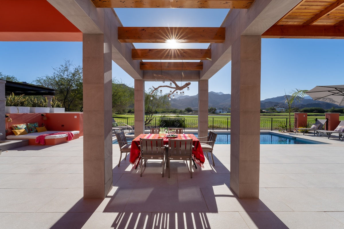 A shaded outdoor dining area is framed by wooden beams, featuring a large table set for meals. The space is surrounded by views of the lush landscape and distant mountains, with a swimming pool visible nearby under clear blue skies.