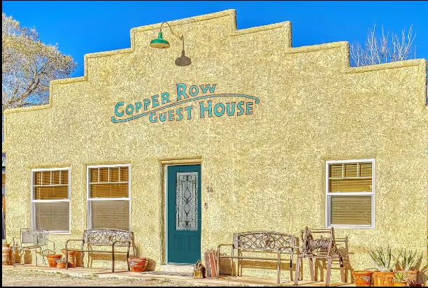The exterior of Copper Row Guest House is showcased, featuring a distinctive adobe-style facade with a sloped roof. A teal front door serves as the focal point, framed by multiple windows. Benches and seating areas line the porch, inviting relaxation and conversation.