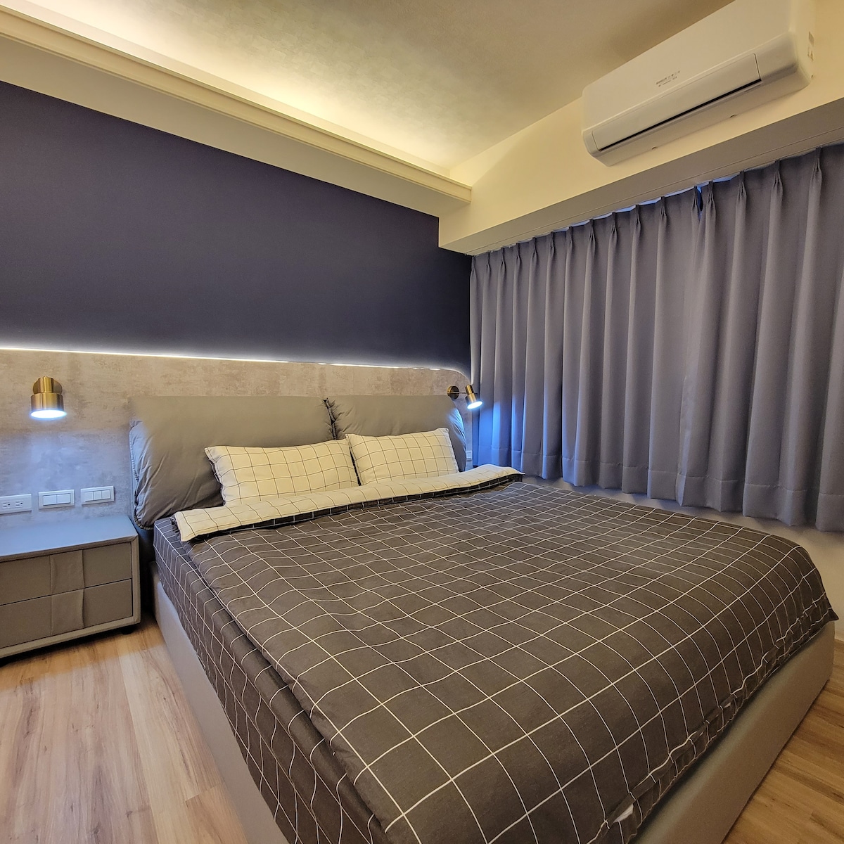 A spacious sleeping area features a large bed with a grid-patterned blanket and soft gray pillows. Beside the bed, modern lamps provide gentle lighting. The wall is accented with dark blue paint, and floor-to-ceiling curtains in a light shade frame the window.
