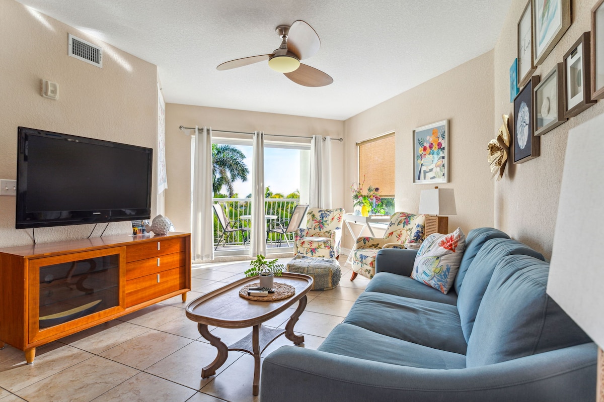 A bright living area is presented with a blue sofa and a wooden coffee table at its center. Light flows through sliding glass doors leading to a balcony. Artwork and plants add interest to the walls, while a television sits on a wooden console.