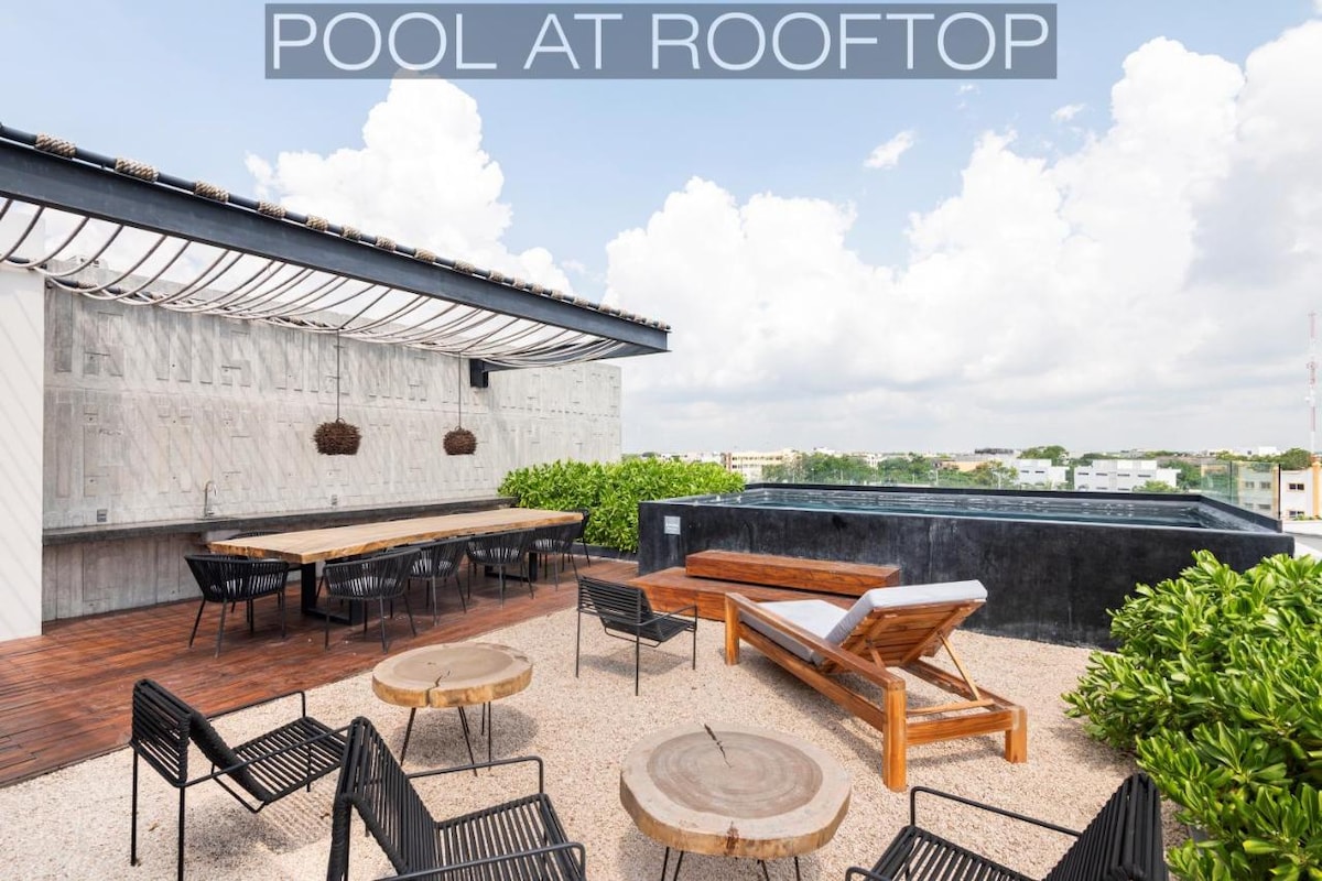 A rooftop space is presented featuring a small pool, surrounded by stylish seating options. Several modern lounge chairs and wooden tables are arranged on a sandy surface, enhanced by green shrubbery and a panoramic view of the surrounding area.