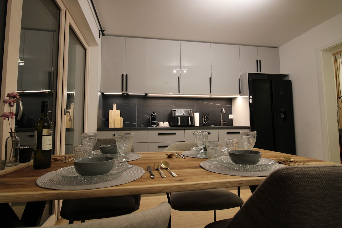 A modern kitchen features sleek gray cabinetry and a dark countertop. A dining table is set with clear glassware and cutlery, accompanied by decorative plates. Natural light enters through large windows, highlighting the clean lines and contemporary design.