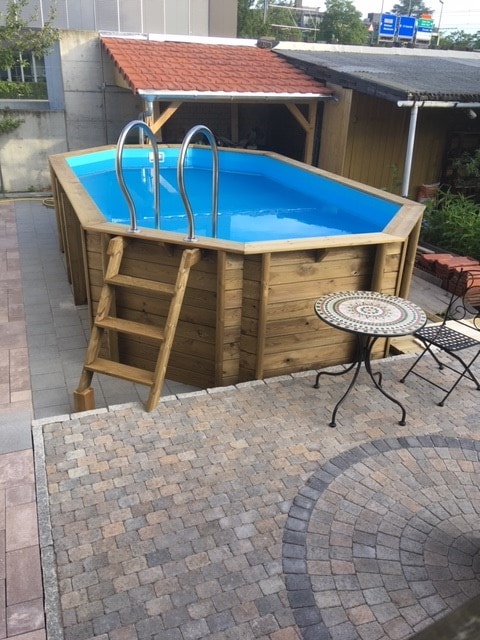 An above-ground wooden pool is featured in the outdoor space, accompanied by a small table and two chairs nearby. A ladder provides access to the pool, while a stone-paved area surrounds it, enhancing the outdoor experience.
