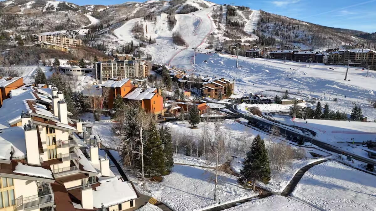 A scenic view captures snow-covered slopes and surrounding accommodations. Towering trees and snowy hillsides provide a serene backdrop. The image showcases the ski area, highlighting accessibility for winter sports enthusiasts, with a clear sky above and a community of cozy buildings below.