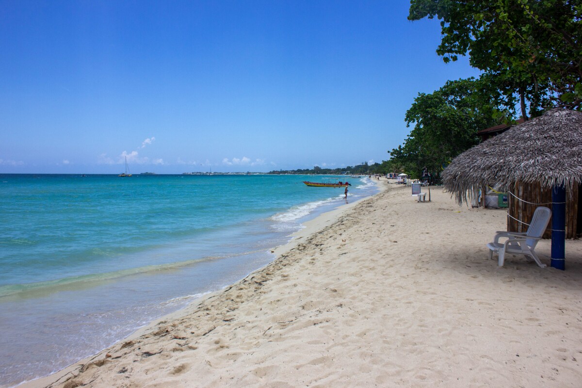 A sandy beach stretches along a serene shoreline, framed by calm turquoise waters. Tall trees provide shade, and local boats are visible in the distance. A thatched-roof hut and sun loungers offer relaxation spots for beachgoers enjoying the inviting landscape.