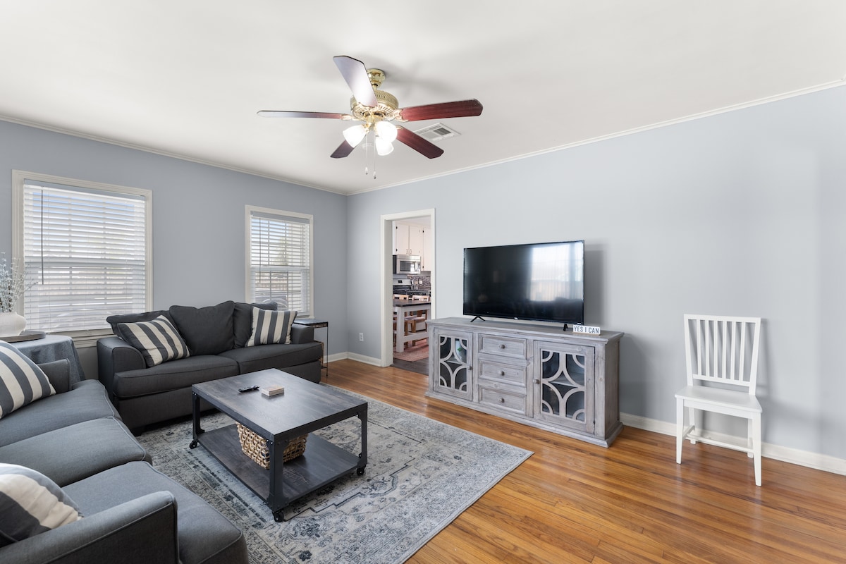 The living area features light blue walls and hardwood flooring. A comfortable sectional sofa is accented by patterned cushions. A coffee table centers the space, with a decorative basket on it. A wall-mounted television is positioned above a gray media console, providing visual balance.