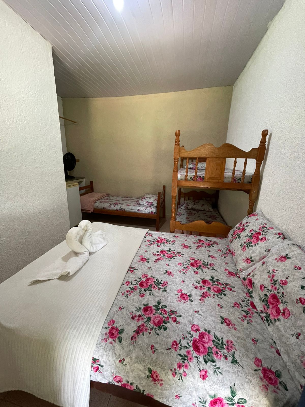 A simple sleeping area reveals two beds, one a spacious single and the other a bunk bed. Floral-patterned bedding adds color to the room. Light from a ceiling fixture illuminates the white walls, creating a bright and inviting space.