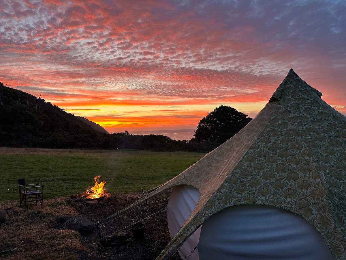 A Lotus Belle tent is positioned beside a fire pit, with a wooden chair nearby. The sky is filled with vibrant sunset colors, casting a warm glow over the expansive lawn and surrounding greenery.
