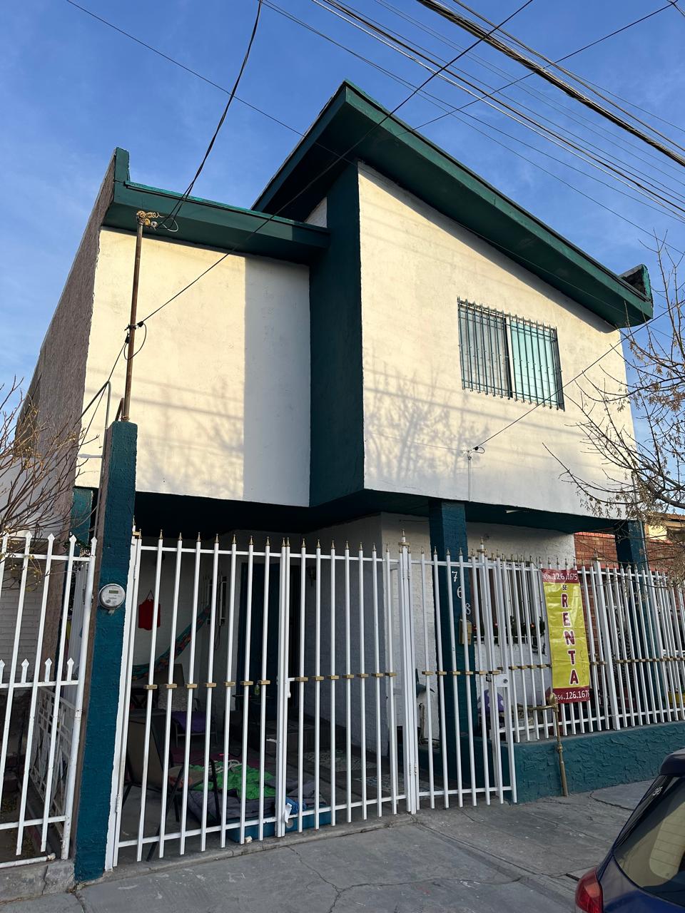 The exterior of a two-story building is shown, featuring a white facade with green trim. A balcony is present above the entrance, and a security fence surrounds the property. The scene is bright, with clear skies and soft shadows at the lower part of the walls.