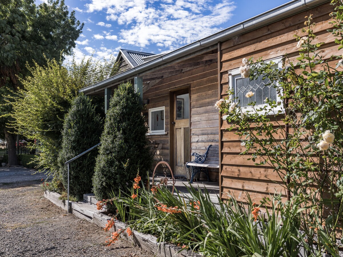 A wooden exterior of the Spud Diggers Hut is framed by lush greenery, including tall hedges and flowering plants. A small, inviting bench is positioned near the entrance, and the sky is dotted with clouds, creating a serene atmosphere around the entrance.