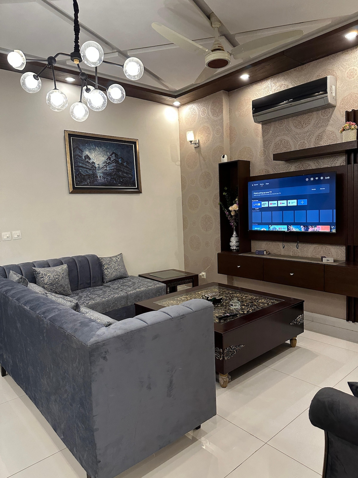 A stylish living area features a plush sectional sofa in muted tones. A dark wood entertainment unit displays a large flat-screen TV. Soft lighting is provided by a unique ceiling fixture, and a wall art piece adds character to the space.