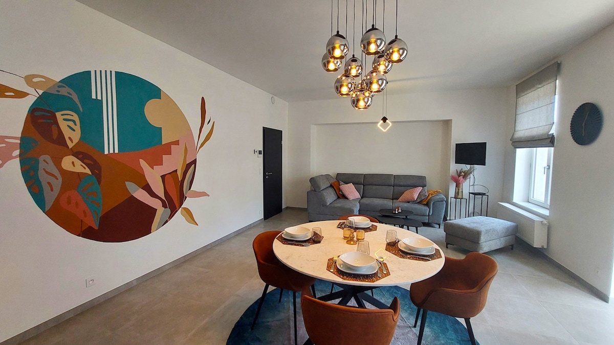 A dining area with a round table set for four features stylish, brown chairs. A large artwork is displayed on the wall, while a comfortable gray sofa is positioned nearby. Bright lighting is provided by a modern chandelier overhead, and a cozy lounge chair complements the seating arrangement.