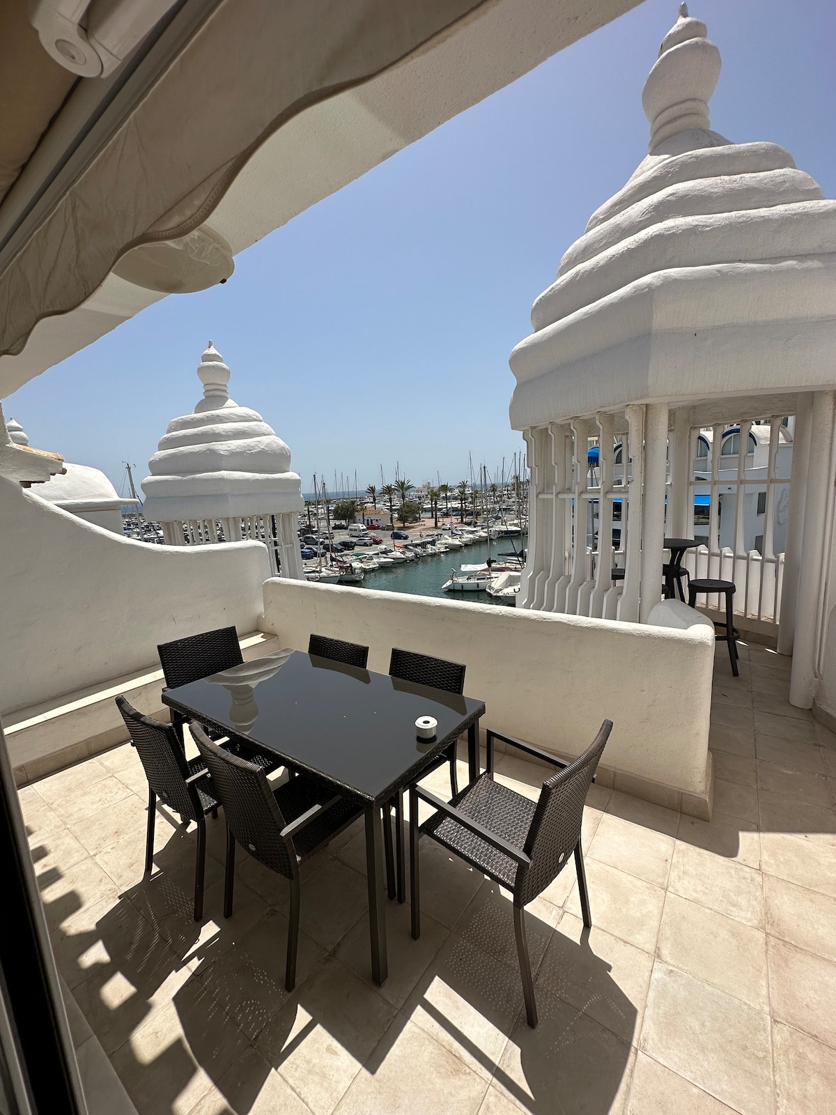 A spacious balcony features a sleek black dining table surrounded by six chairs, offering a view of the marina. The area is adorned with decorative white structures reaching towards the sky, with bright sunlight illuminating the scene.