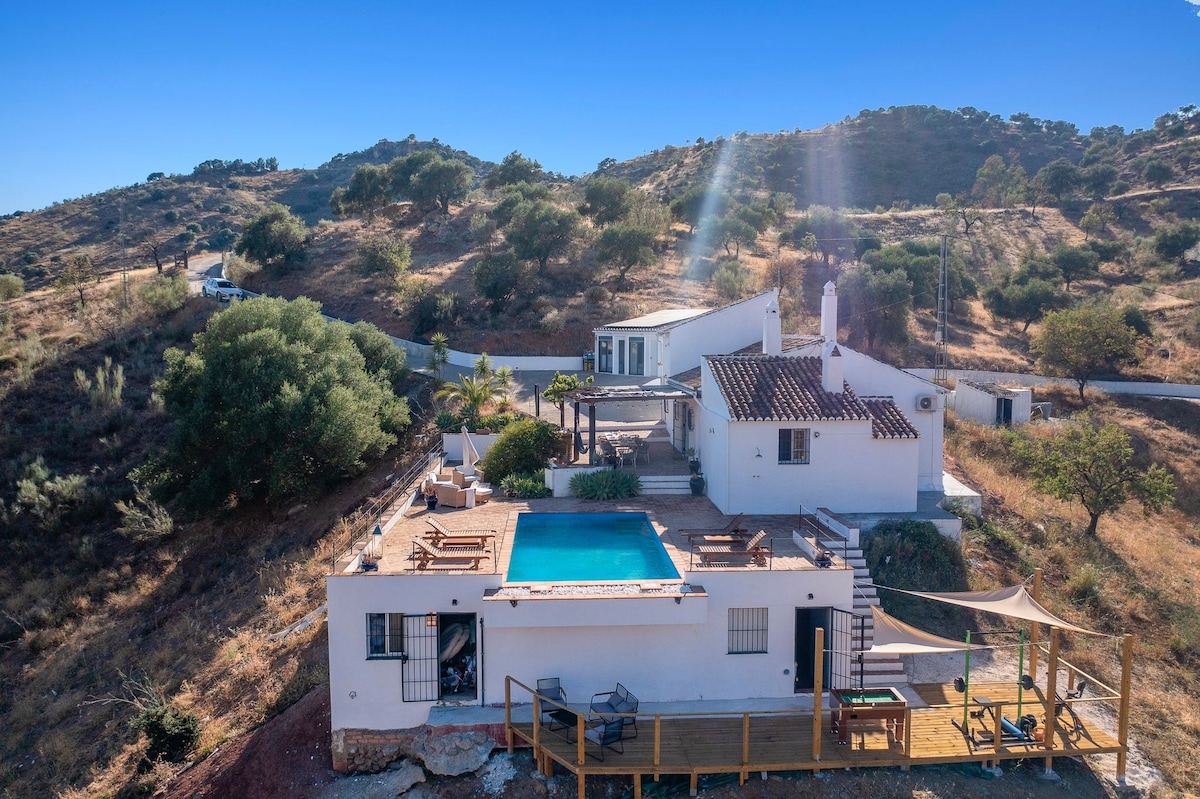 A spacious retreat is situated on a hillside, surrounded by nature. The image captures a private pool framed by sun loungers and shaded areas. The white exterior of the house contrasts with the earthy tones of the landscape, showcasing a relaxing outdoor space perfect for gatherings.