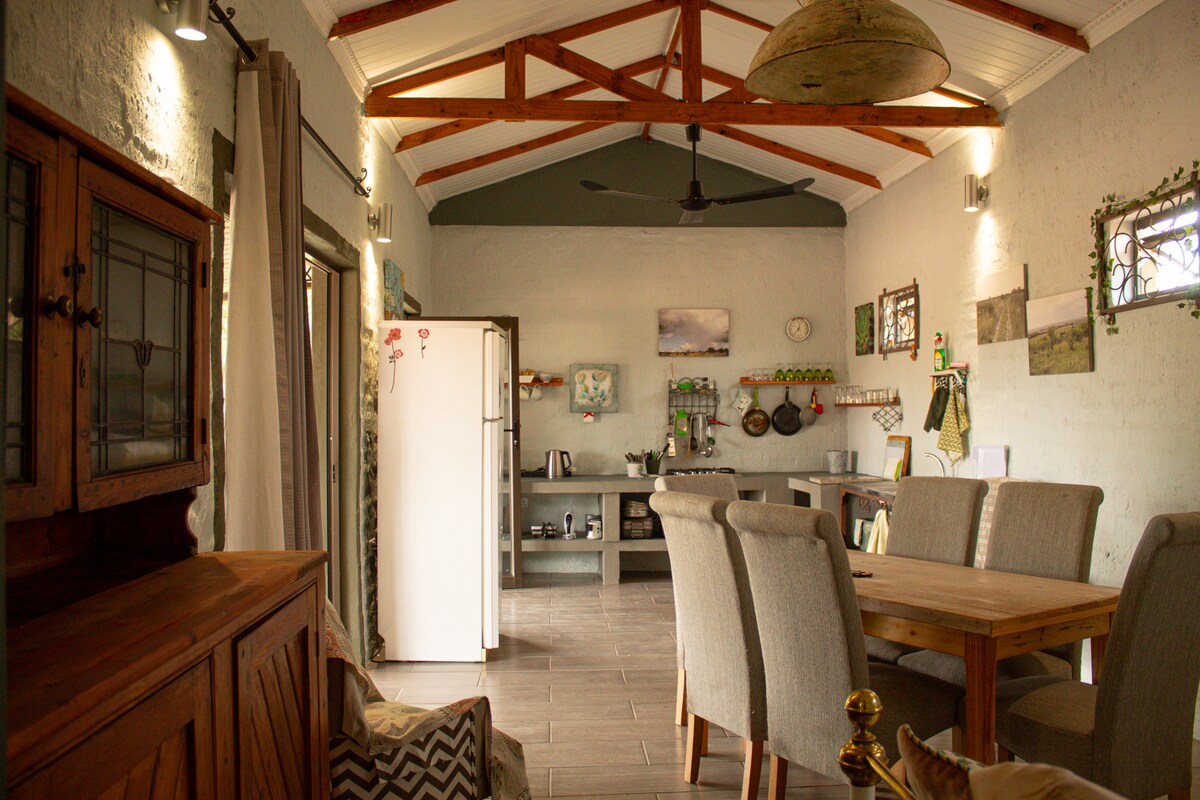 A spacious living area features a wooden dining table surrounded by upholstered chairs. A refrigerator and various kitchen items are visible along one wall, complemented by decorative artwork and plants that enhance the setting's natural feel.