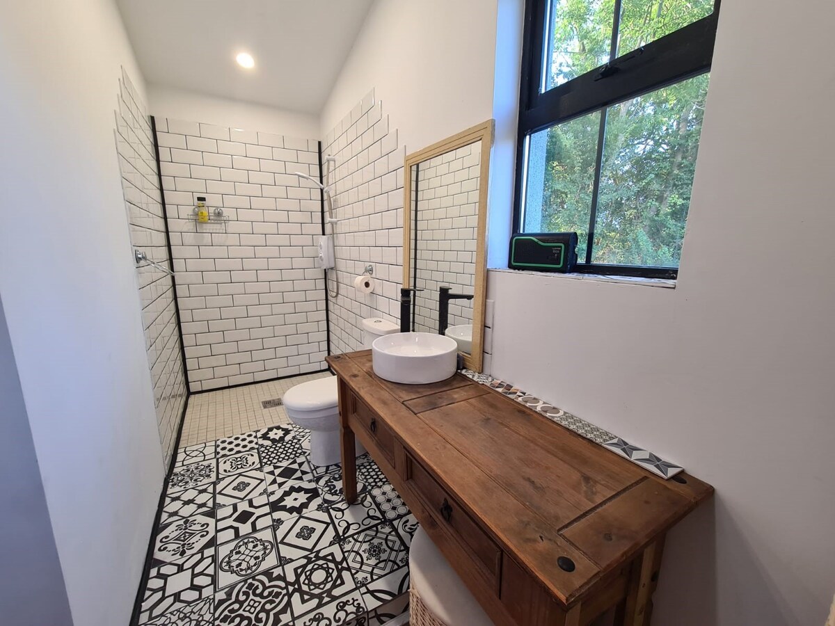 A spacious bathroom is highlighted by white tiled walls and a contrasting patterned floor. A modern round sink is placed atop a wooden vanity, with a large mirror mounted above. Natural light filters through a window, enhancing the bright atmosphere.