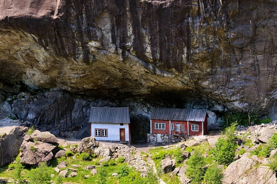 Two rustic cabins are situated at the base of a rocky cliff, surrounded by greenery and boulders. One cabin features a red exterior, while the other is painted white. The natural landscape presents a sense of seclusion and tranquility under the imposing stone formation.