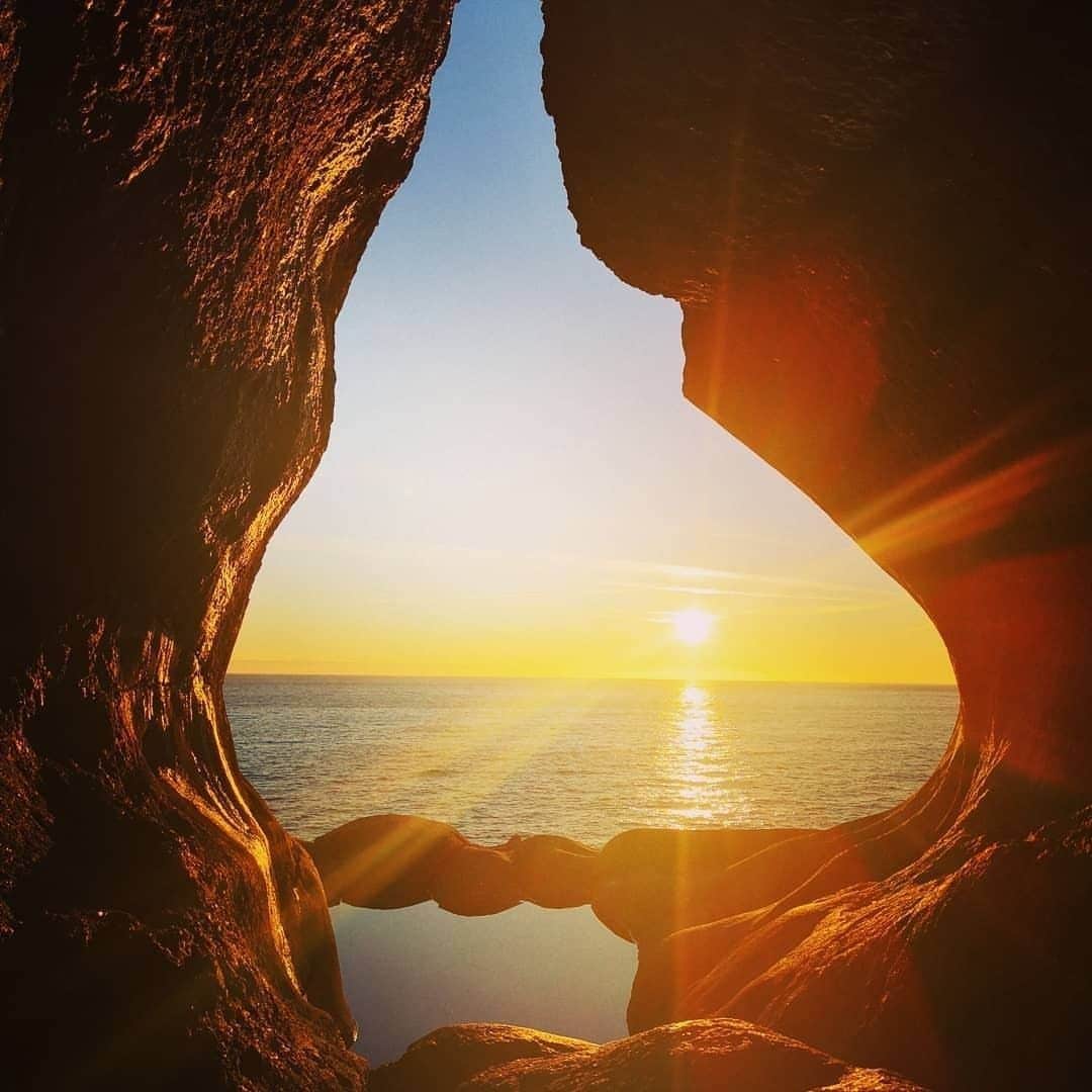 Sunlight filters through rock formations, illuminating the scene. A calm sea is visible outside, reflecting the golden hues of a setting sun. The framing stone creates a natural arch, enhancing the scenic view.