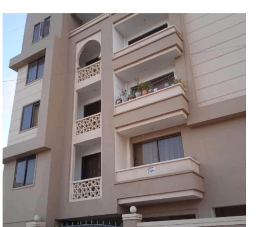The exterior of a multi-story building is illustrated, featuring a modern façade in neutral tones. Balconies with decorative railings are visible, adorned with potted plants. Large windows allow natural light to enter, and a welcoming entryway is framed by a decorative arch.