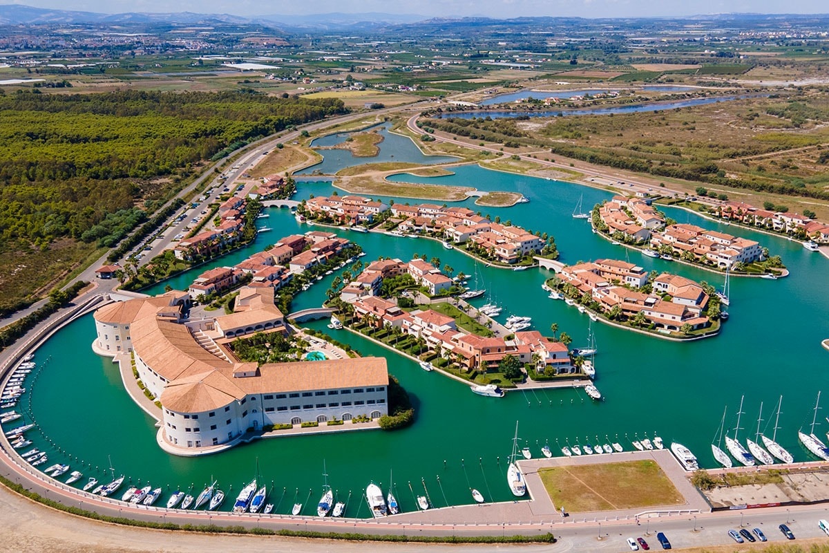 An aerial view of a serene marina complex showcases an array of interconnected buildings surrounded by lush greenery. Calm turquoise waters reflect the architecture, while numerous boats are docked along the waterfront. Scenic pathways and landscape contribute to the tranquil ambiance of the area.