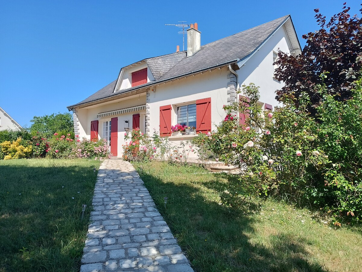 A charming house is set against a clear blue sky, showcasing a well-maintained garden with blooming flowers. A stone pathway leads to the front door, framed by red shutters. The structure features a welcoming porch and a green lawn, adding to its inviting appearance.