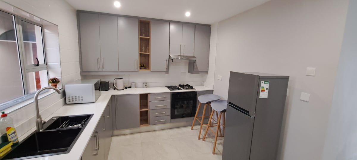 The kitchen features modern cabinetry in neutral tones, with sleek countertops and stainless steel appliances. A double sink sits under a large window, while a refrigerator and stove are positioned for convenience. Two high-backed stools are arranged at a small breakfast bar.