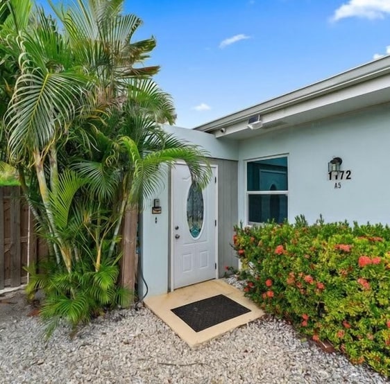 The entrance is framed by lush tropical plants and vibrant flowers, creating an inviting approach to the home. A decorative door with a glass panel is positioned to the right of the image, with a welcome mat in front, suggesting a warm and friendly atmosphere.
