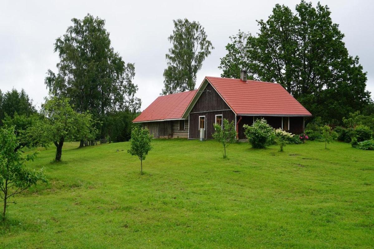 The exterior of a charming wooden house is set amid lush greenery. A red-tiled roof contrasts with the natural surroundings. Large trees and various shrubs are visible, adding to the tranquil atmosphere of the landscape.