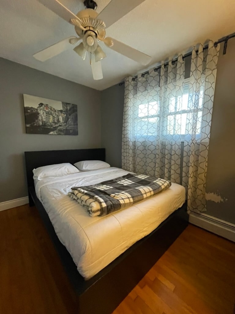 A comfortable bedroom features a dark wooden bed with a neatly arranged white duvet and a plaid throw blanket. Two windows provide soft natural light, framed by patterned curtains. A piece of artwork hangs on the wall, contributing to the room's calming atmosphere.