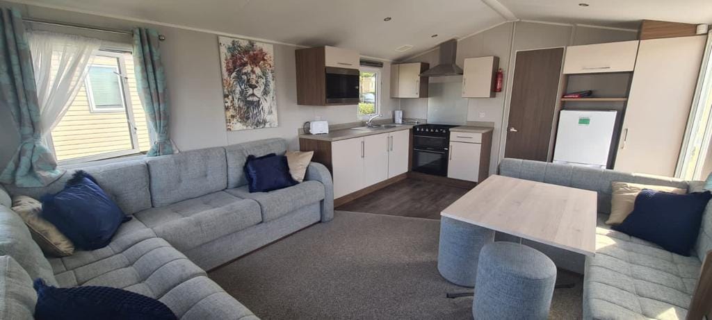 A modern living area features comfortable grey sofas arranged around a light wood coffee table. Natural light enters through large windows adorned with soft curtains. A well-equipped kitchen with sleek cabinetry and appliances can be seen in the background, enhancing the open layout.