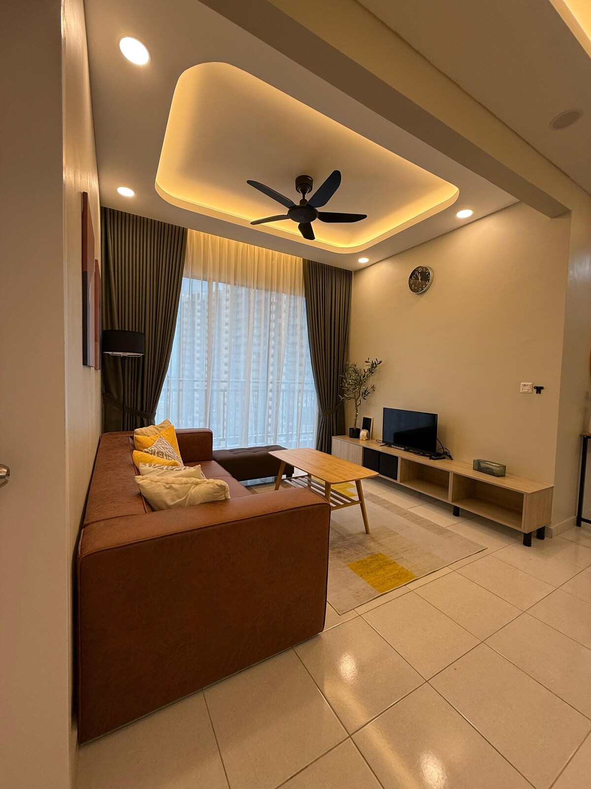 A modern living area is illuminated by soft ceiling lights. A cozy brown sofa is complemented by a coffee table. A television is positioned against the wall, with a large window draped in sheer curtains allowing natural light to fill the space.