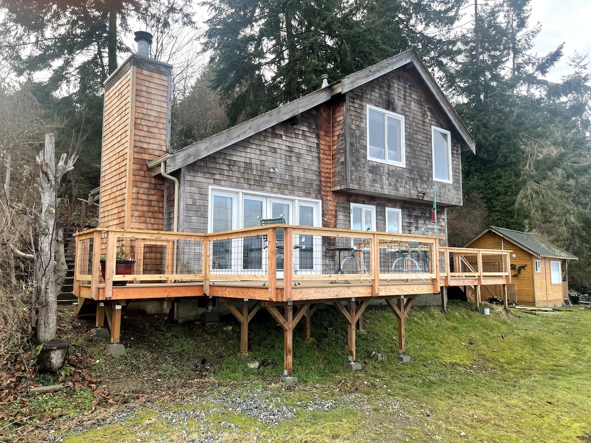 The wooden cottage features a spacious front deck, providing an outdoor seating area with views of the surrounding nature. The structure is accented by a chimney and large windows that allow for ample natural light. The nearby yellow storage shed can be seen on the property.
