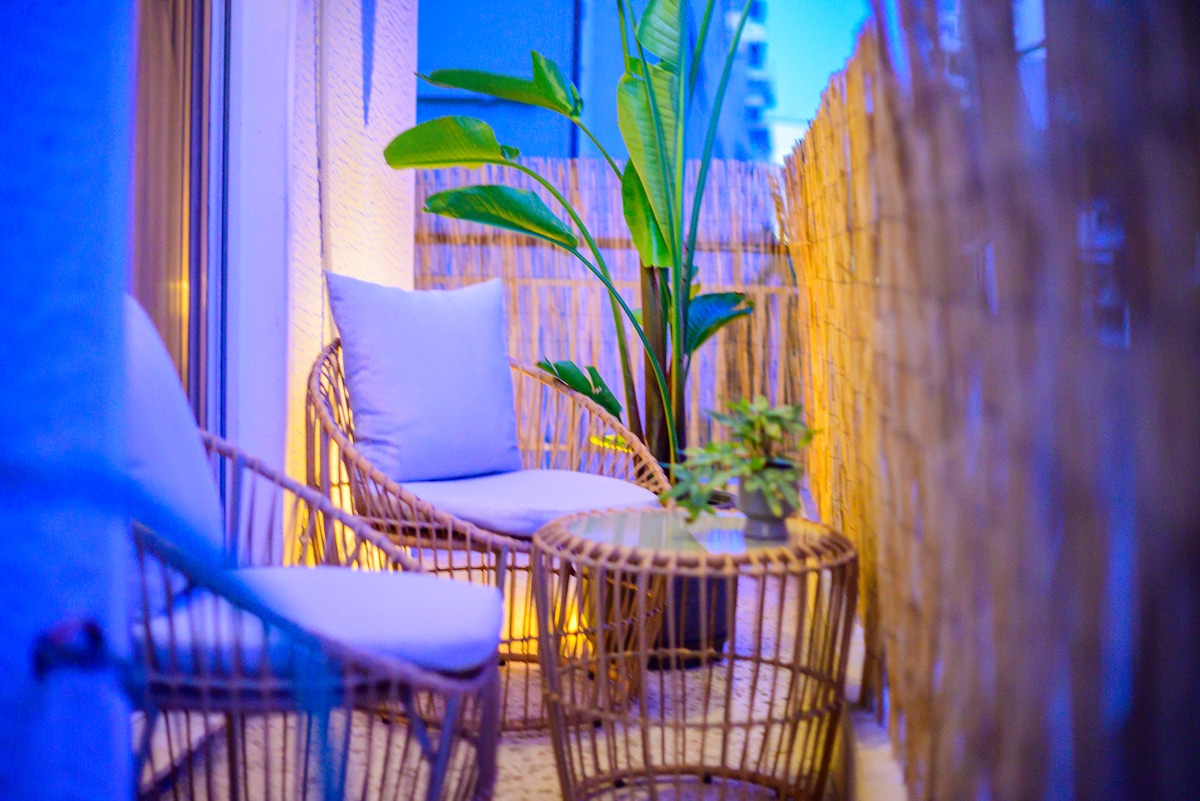 A cozy balcony is featured, furnished with two rattan chairs and a small table. Soft blue lighting creates a calm ambiance, while a tall green plant adds a touch of nature. A bamboo fence provides privacy, enhancing the space's intimate feel.