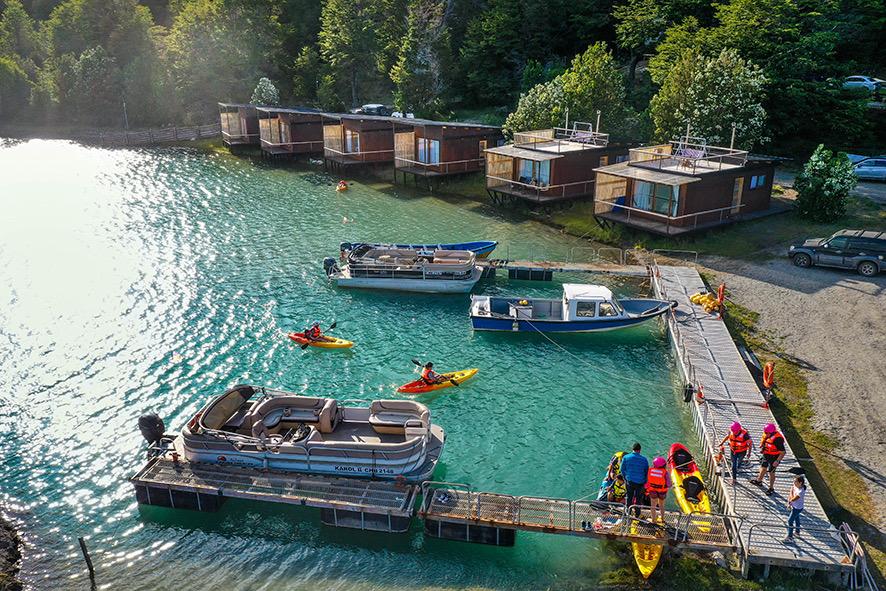 A serene waterfront scene features several wooden cabins by a clear blue lake. Kayaks are being launched from a dock, where a group of individuals are seen gathering near boats. Lush greenery borders the area, creating a harmonious connection with nature.