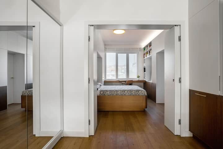 A bright and cozy bedroom featuring a large window, a comfortable double bed with floral bedding, and ample built-in storage. Perfect for relaxation after a long day exploring the city.