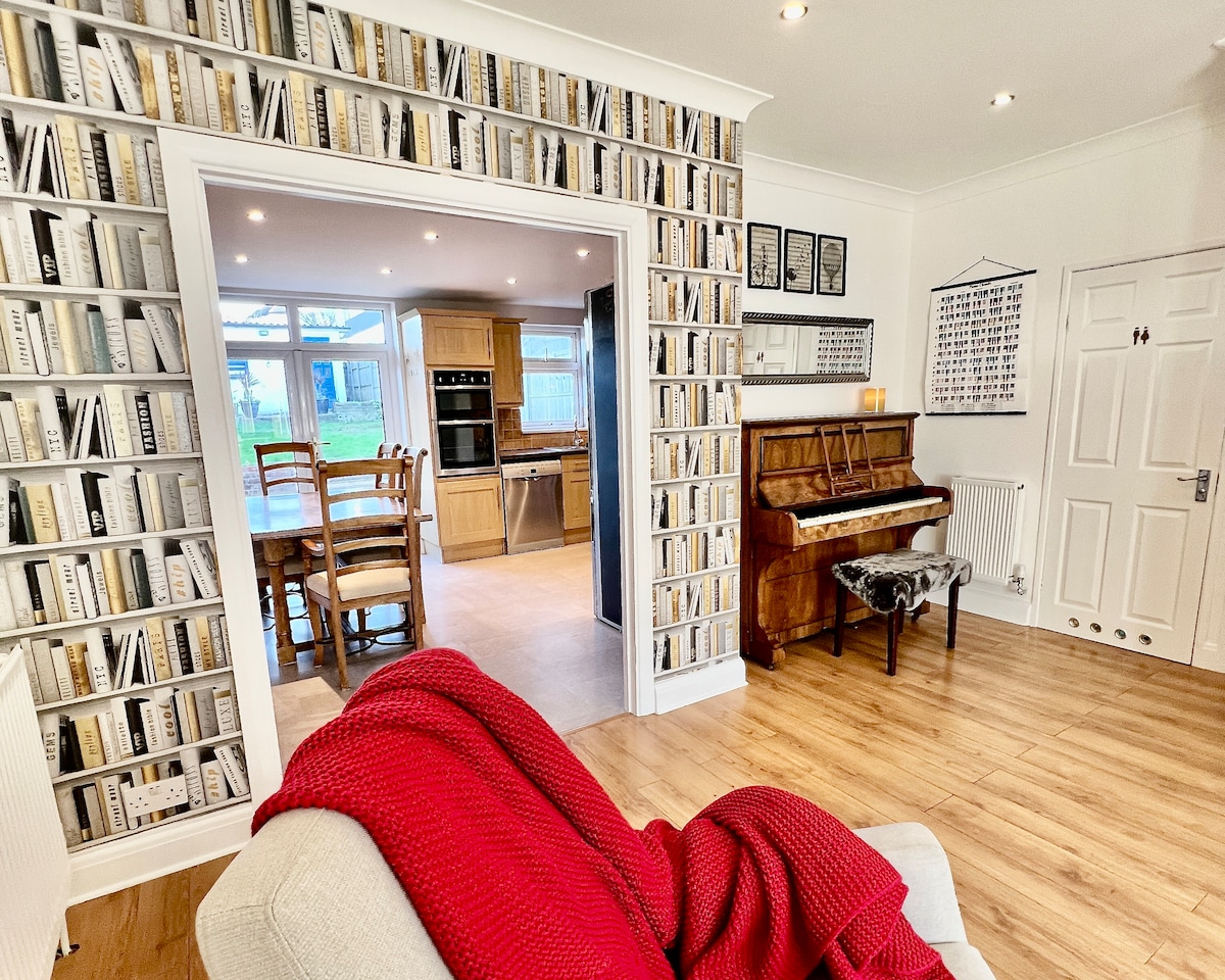A cozy living area features a light-colored sofa with a textured red throw. A piano is situated near bookshelves wallpaper, and a dining space can be seen through an archway, where wooden furniture complements the modern kitchen in the background.