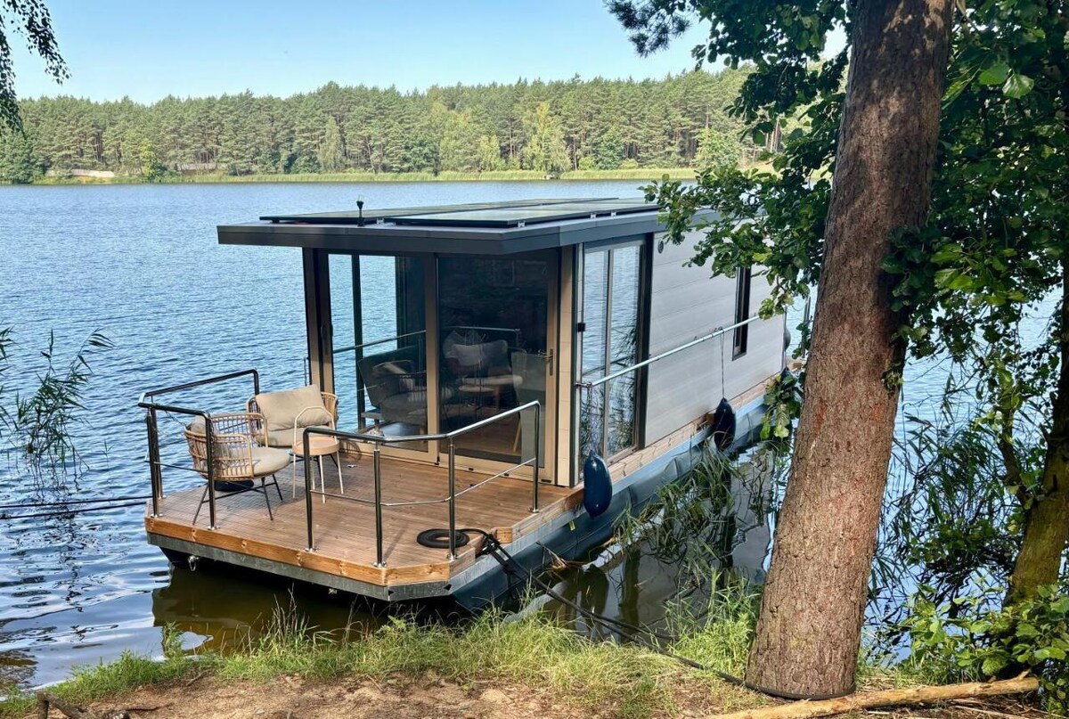A houseboat is anchored at the edge of a serene lake, surrounded by lush greenery. Large windows frame the view of the water, and a small deck features outdoor seating arrangements, inviting relaxation amidst nature.