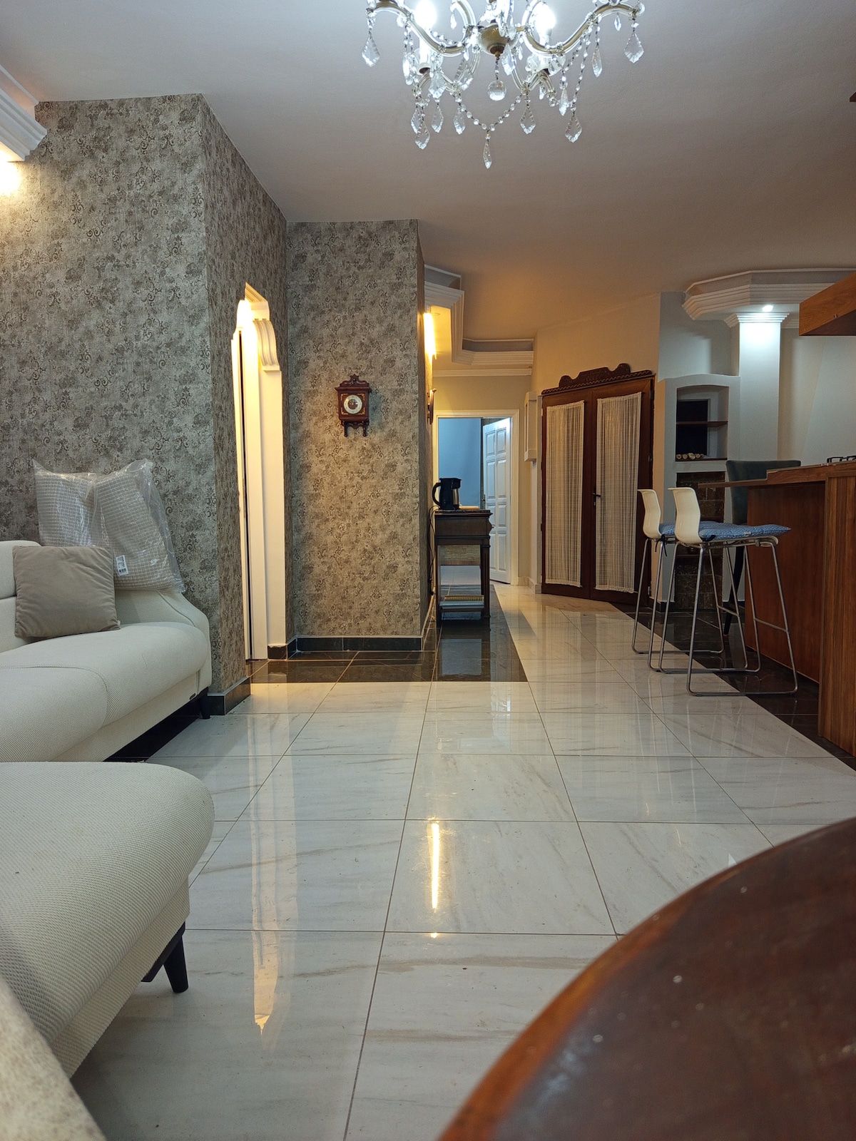 A spacious living area is viewed that features a light-colored sofa and a wooden dining table. The floor is tiled in a glossy, neutral tone. A chandelier adds subtle elegance, while the nearby kitchenette is partially visible, showcasing a modern setup.