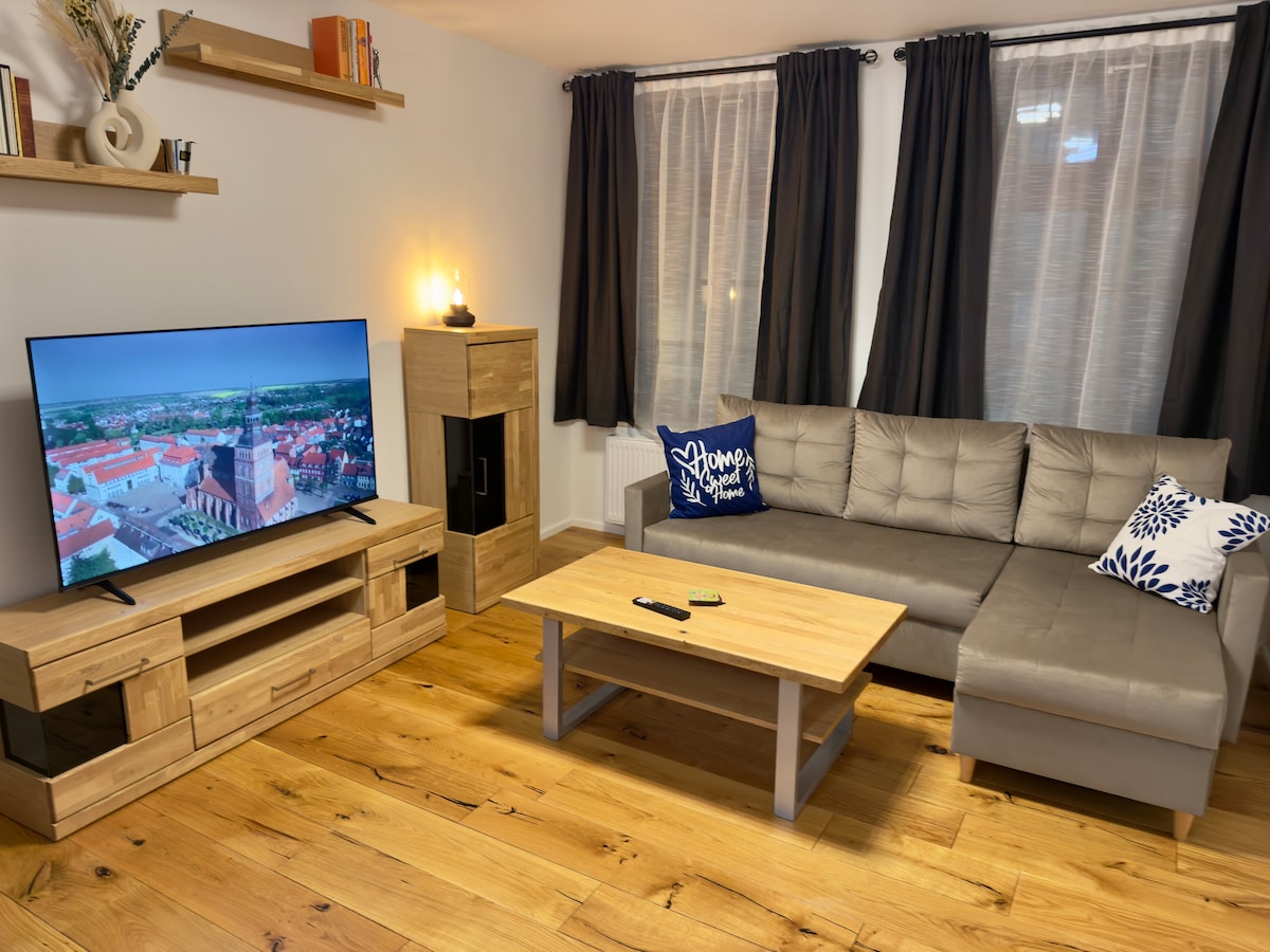 A welcoming living area is presented, featuring a comfortable sectional sofa and a wooden coffee table. A television is positioned on an entertainment unit, complemented by a decorative piece. Large windows, framed by dark curtains, allow natural light to fill the space, enhancing the wooden floor's warmth.