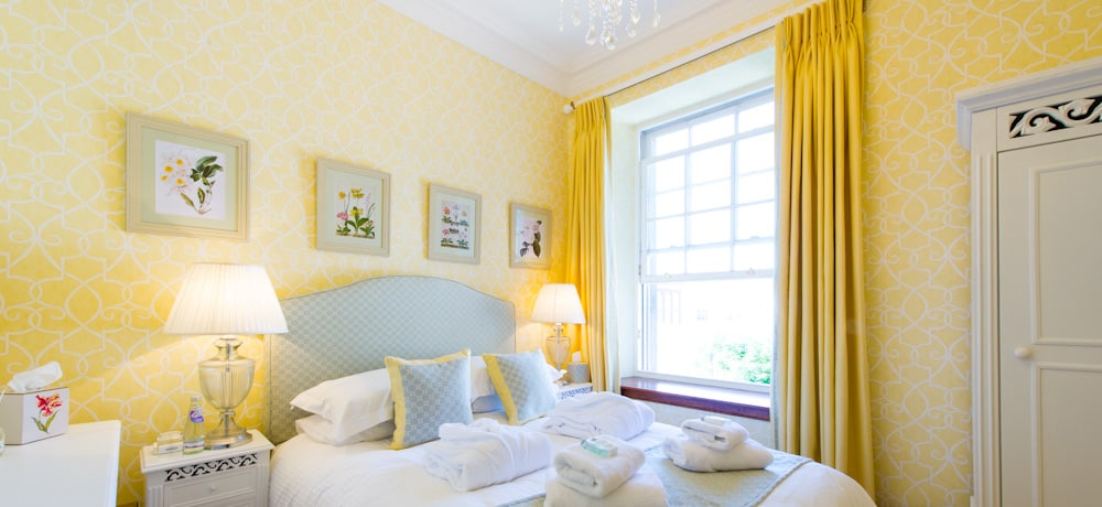 A bright bedroom is adorned with cheerful yellow wallpaper and framed botanical prints. The spacious bed is dressed in crisp linens, while fluffy towels are neatly arranged on the bed. Natural light filters through a large window, complemented by soft yellow curtains that enhance the room's light ambiance.