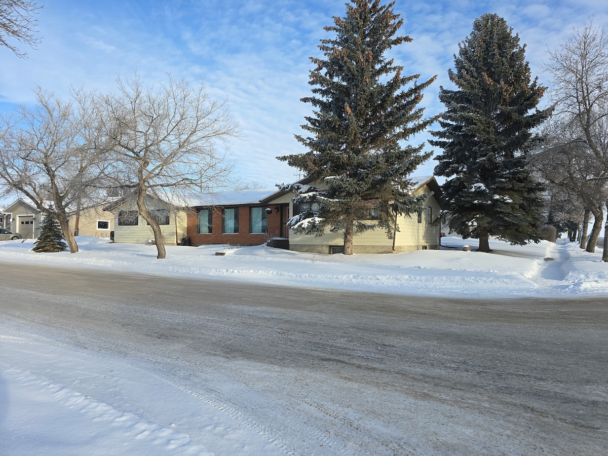 A large house is positioned on a snowy street, surrounded by tall evergreen trees. The exterior features a combination of brick and siding, with large windows allowing for natural light. Snow blankets the ground, creating a serene winter scene.