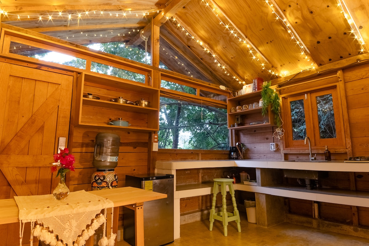 The kitchen area features wooden walls and shelving, illuminated by soft string lights along the peaked ceiling. A small table is positioned beside a mini-fridge and a sink, with a gas stove placed on the countertop. A water dispenser is visible, enhancing the functional appeal of the space.