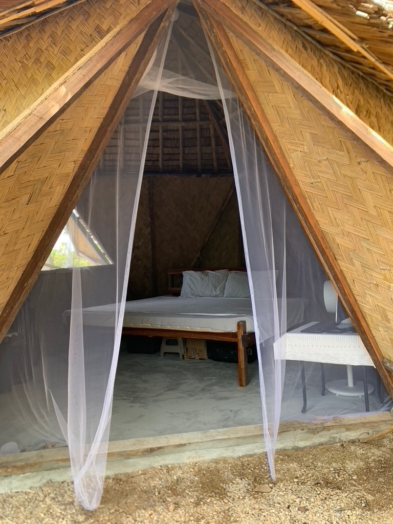 An entrance to a dome-shaped structure is framed by natural bamboo walls, showcasing a spacious interior. A bed is visible, draped with a protective mosquito net. A simple chair sits nearby, and soft natural light filters through the open doorway.